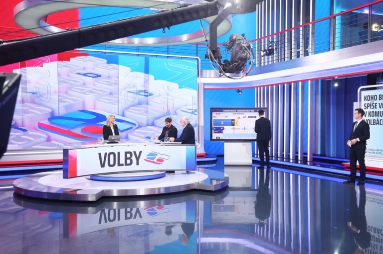 Televizní studio volebního vysílání, moderátorka a hosté u stolu, další moderátor u obrazovky.