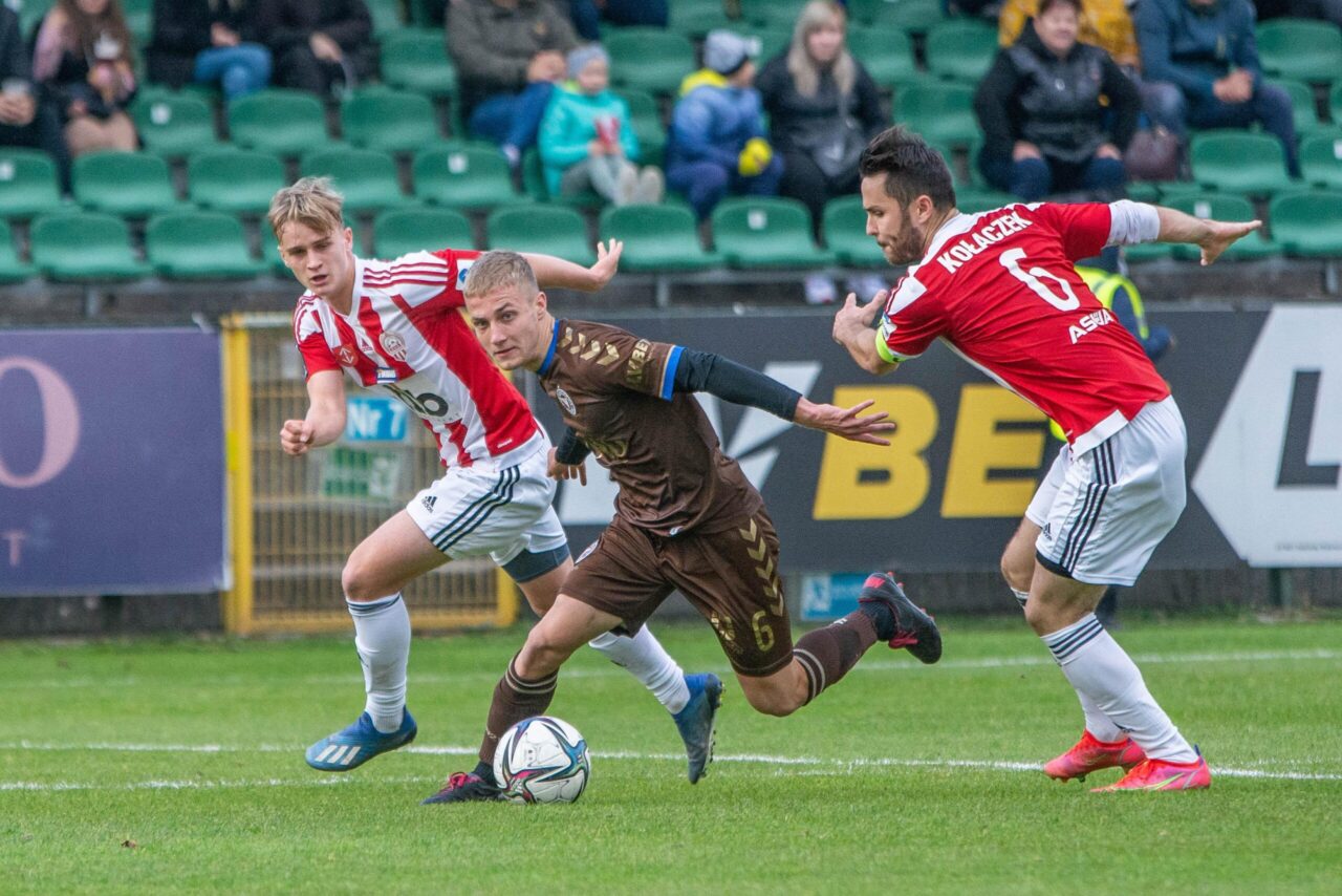 Fotbalista v hnědém dresu vede míč, dva protihráči v červenobílém ho brání.